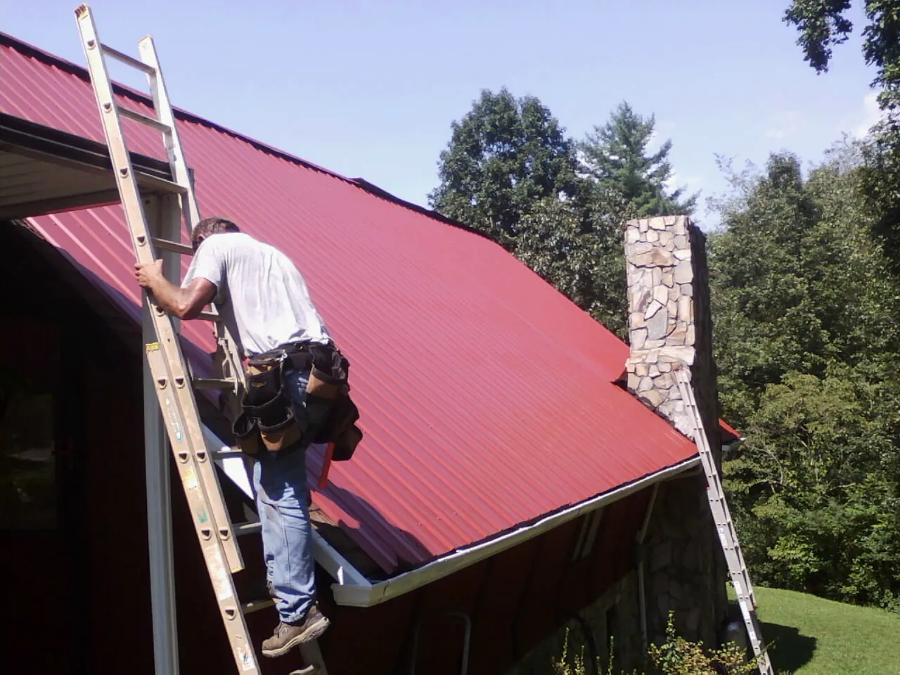 Roofer on a ladder beside a red metal roof and stone chimney — Soffit & Fascia Repair by Roofing Brothers in Maple Falls